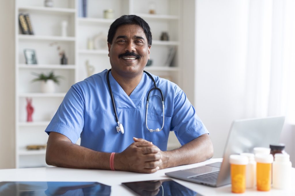 Smiling Indian Male Doctor In Blue Uniform Sitting At Desk With Laptop